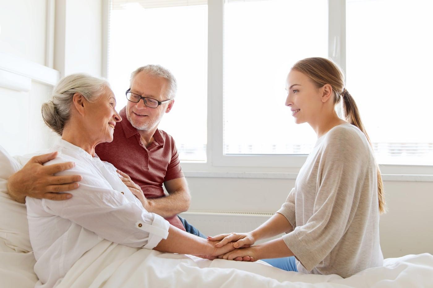 Family members holding hands with an older woman in a hospital bed